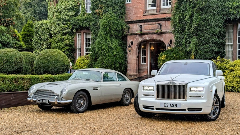 A white Rolls-Royce Phantom parked next to a classic Aston Martin DB5 in front of a country house wedding venue.